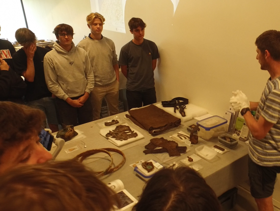 We zien een groep leerlingen rond een tafel vol archeologische vondsten staan. Jan Decorte, archeoloog bij CO7 geeft uitleg. 