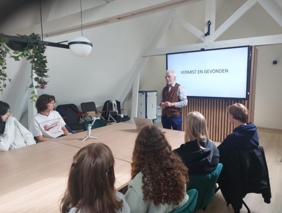 Een groep leerlingen zit aan tafel en luistert naar Pol Lefevre die aan het hoofd van de tafel staat. Achter hem staat een scherm met de beginslide van zijn presentatie. 